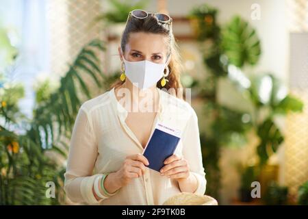 Reise während der Coronavirus-Pandemie. Trendige Touristenfrau mit Covid-Pass, medizinischer Maske, Flugticket und Sonnenbrille im modernen Zuhause an sonnigen Tagen. Stockfoto