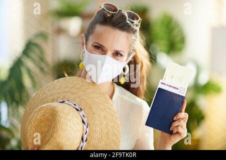 Reisen während der Covid-19-Pandemie. Porträt einer trendigen Reisenden Frau mit covid Pass, medizinische Maske, Flugticket, Strohhut und Sonnenbrille in der mod Stockfoto