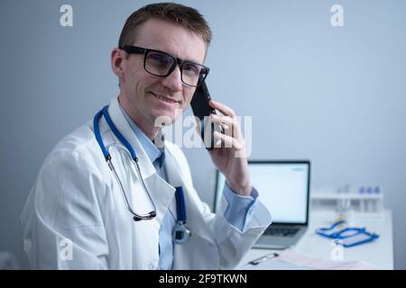 Europäischer Arzt in weißem medizinischen Mantel und Brille konsultiert den Patienten auf dem Handy in der Klinik Büro, sitzen am Tisch mit Laptop. Deutscher General Stockfoto