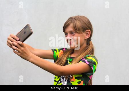 Ein zehn Jahre altes Mädchen posiert und macht ein Selfie mit der Hand auf ihrem Handy in einem braunen Fall. Grauweißer Hintergrund. Lächelnd attraktives Mädchen hat Spaß Stockfoto