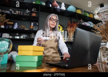 Shopping- und Discountsystemkonzept. Sympathische grauhaarige Dame, die auf einem modernen Laptop tippt, während sie mit einer Rabattkarte an der Theke sitzt. Regale mit verschiedenen schönen Dekor auf Hintergrund. Stockfoto