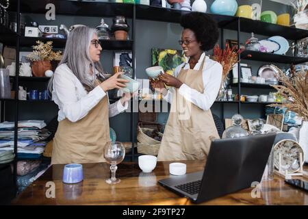 Kaukasisch gealterte Frau und afrikanische junge Frau, die gemeinsam Inventar in einem modernen Geschäft mit verschiedenen Dekorationen machen. Zwei verschiedene Mitarbeiter, die im Geschäft einen drahtlosen Laptop verwenden Stockfoto