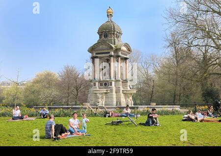 Junge Leute, die im Frühling im Sarphatipark auf Gras relaxen, mit Monument Samuel Sarphati im Hintergrund, De Pijp, Amsterdam, Niederlande, Europa Stockfoto