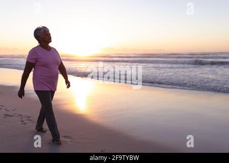 Ältere afroamerikanische Frau, die während des Sonnenuntergangs am Strand spazieren geht Stockfoto