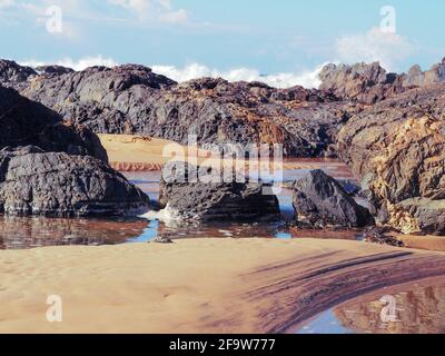 Abstrakte Strandszene an der Küste, wunderschöne warme rote Brauntöne mit weitläufigen Linien, natürliche Muster im Sand, Felsenbecken mit krachenden Meereswellen Stockfoto