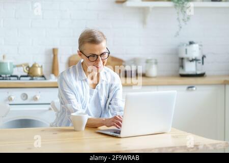 Moderne, glückliche, reife Frau, die eine Brille trägt, die am Laptop arbeitet oder studiert, während sie zu Hause in der Küche sitzt und auf den Bildschirm schaut und etwas auf der Tastatur tippt. Konzept für Führungskräfte und Technologien Stockfoto