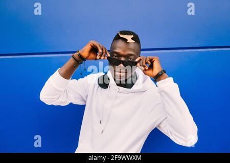 Ein junger afroamerikanischer schwarzer Mann, der ein weißes Sweatshirt, eine Sonnenbrille, einen Kamm und Kopfhörer an einer blauen Wand trägt und voller Zuversicht über die Brille blickt Stockfoto