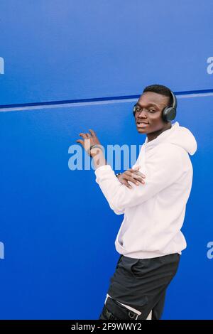 Ein junger afroamerikanischer schwarzer Mann, der ein weißes Sweatshirt und Kopfhörer trägt, geht an einer blauen Wand entlang und schaut auf die Kamera. Moderner Look Stockfoto