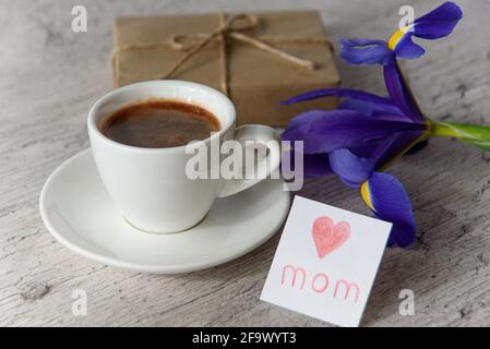 Weiße Tasse Kaffee, Karte für Mama vom Kind, Geschenkbox und Irisblume auf grauem Holztisch. Alles Gute Zum Muttertag Stockfoto