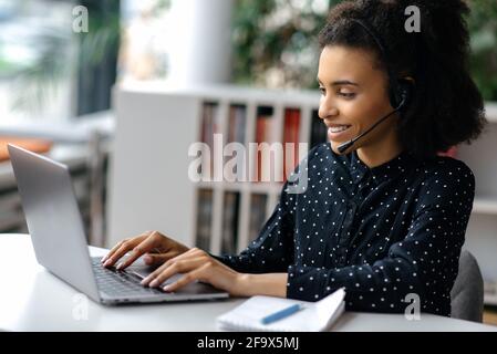 Afroamerikanisch schöne junge Frau, die ein Headset trägt, am Schreibtisch sitzt, einen Laptop benutzt, online arbeitet, im Internet surft, mit Kollegen oder Kunden chattet, E-Mails beantwortet und lächelt Stockfoto