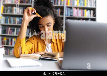 Eine intelligente, auf afroamerikanische millennials fokussierte Studentin, die mit einem Laptop an einem Tisch in der Universitätsbibliothek sitzt und mit Interesse Buch liest, sich auf Vorlesung oder Prüfung, Wissen und Lernkonzept vorbereitet Stockfoto