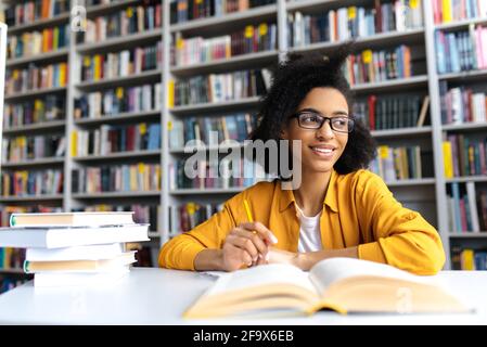 Teenager afroamerikanische hübsche Schülerin mit Brille, studiert, während sie am Tisch in einer Universitätsbibliothek sitzt, Bücher liest, um Informationen für eine Lektion oder Prüfung zu suchen, Hausaufgaben macht, schaut weg Stockfoto