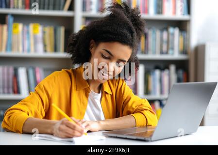 Eine fröhliche afroamerikanische, smarte Studentin mit lockigen Haaren, trendig gekleidet, sitzt mit einem Laptop an einem Schreibtisch in der Universitätsbibliothek, fokussiert während eines Vortrags Notizen machen und lächeln Stockfoto