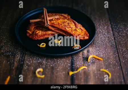 Zwei Scheiben gebratenes Brot mit zwei Stäbchen Zimt, Zucker und Zitronen- und Orangenschalen auf einem schwarzen Teller auf einem dunklen Holztisch Stockfoto