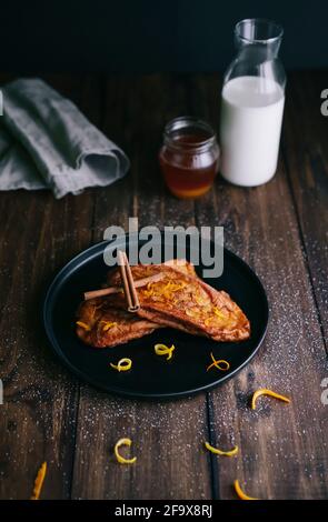 Gebratene Brotscheiben mit zwei Zimtschnecken, Zucker und Orangen- und Zitronenschalen auf einem schwarzen Teller auf einem dunklen Holztisch Stockfoto