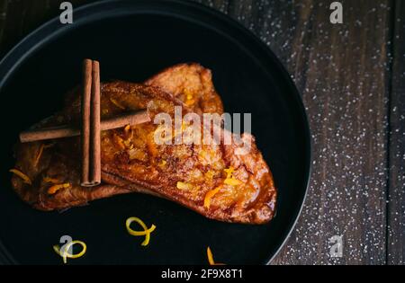 Luftaufnahme von zwei Scheiben gebratenes Brot mit Honig, Zucker, Orangen- und Zitronenschalen und zwei Zimtstangen auf einem schwarzen Teller auf einem dunklen Holztisch Stockfoto