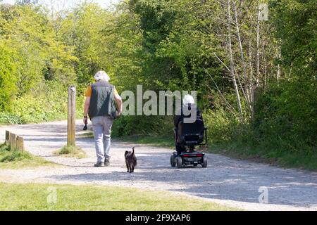 Ein Paar mittleren Alters auf dem Land, das mit dem Hund unterwegs ist Mit der Dame, die einen Mobilitätsroller benutzt Stockfoto