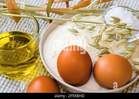 Lebensmittelzutaten zum Backen, Mehl, Eier, Öl und Maisähren auf weißem Tischtuch auf dem Tisch. Stockfoto