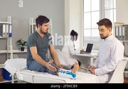 Besorgter junger Mann mit Fußverletzung, der den Arzt im Sitzen konsultiert Untersuchungsbett in der Klinik Stockfoto