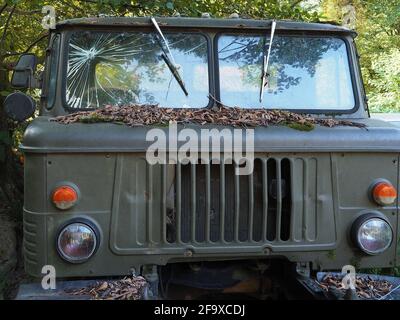 Ein alter verlassener Militärwagen mit zerbrochenem Glas steht verstreut Mit trockenem Laub umgeben von grünen Bäumen Stockfoto