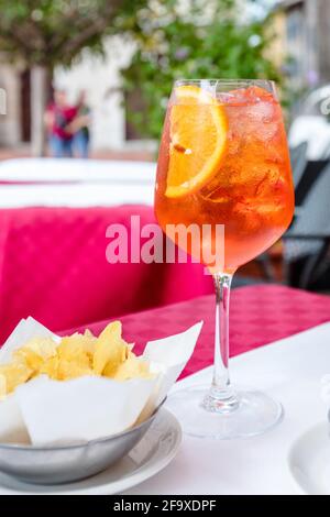 Ein Glas aperol Spritz Cocktail auf einem Tisch im Freien bei Sonnenschein Tag mit einer Schüssel Kartoffelchips im Vordergrund Stockfoto