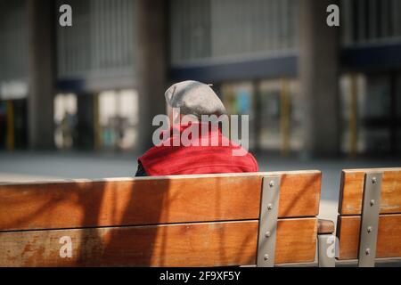 Ein älterer Mann mit einem roten Oberteil und einer grauen Baskenmütze sitzt auf einer Bank vor dem Busbahnhof Preston in der Sonne. Stockfoto