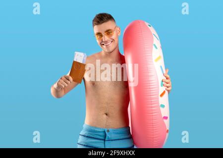 Hübscher junger Mann mit aufblasbarem Ring, der Pass mit Tickets zeigt und auf blauem Hintergrund tropische Ferien macht Stockfoto