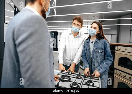 Shop Assistant im Hypermarkt zeigt dem jungen Paar ein neues Modell des Gasofens, das alle medizinische Masken trägt Stockfoto