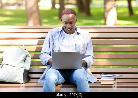 Aufgeregter schwarzer Student, der auf den Laptop-Bildschirm schaut, kann seinen Erfolg im Park nicht glauben Stockfoto