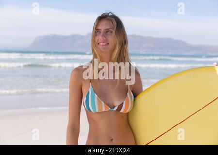 Lächelnde kaukasische Frau trägt Bikini mit gelbem Surfbrett an der Strand Stockfoto