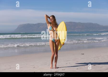 Lächelnde kaukasische Frau trägt Bikini mit gelbem Surfbrett an der Strand Stockfoto