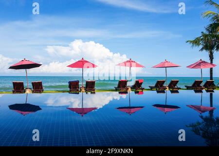 Tropisches Resort mit Pool in der Nähe von Palmenstrand an einem sonnigen Tag, Sonnenliege und rotem Sonnenschirm in der Nähe des Meeres. Stockfoto