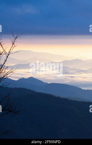 Landschaft mit blauen Bergketten im Morgennebel, dramatischen blauen Wolken über den Bergen, einem Dorf und einem Fluss im nebligen Tal. Thailand. Stockfoto