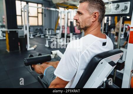 Attraktiver, selbstbewusster, gesunder Sportmann mit sitzender Beincurl-Maschine im Fitnessstudio, Seitenansicht Stockfoto