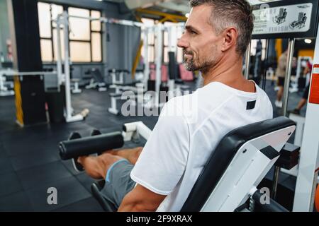 Attraktiver, selbstbewusster, gesunder Sportmann mit sitzender Beincurl-Maschine im Fitnessstudio, Seitenansicht Stockfoto