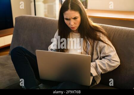 Unglücklich gestresste Frau, die auf den Laptop-Bildschirm schaut und zu Hause auf einer Couch sitzt Stockfoto