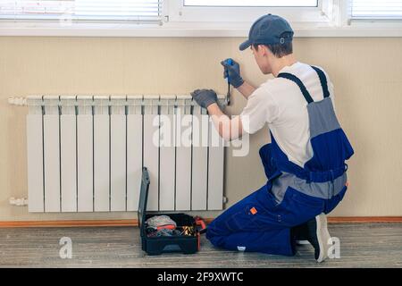 Ein Klempner in einem blauen Overall fixiert einen Kühler. Mit einem verstellbaren Schraubenschlüssel zieht er den Hahn fest. Eine Aktentasche mit einem Werkzeug zur Hand Stockfoto