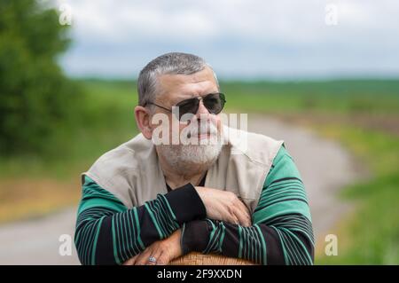 Schönes Outdoor-Porträt von bärtigen kaukasischen älteren Mann in schwarz Sonnenbrillen lehnen sich auf einen Korbsessel Stockfoto
