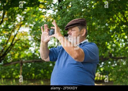 Kaukasischer bärtiger älterer Mann, der im Freien ein Mobiltelefon verwendet, um Fotos zu machen Mit interner Kamera in der Sommersaison Stockfoto