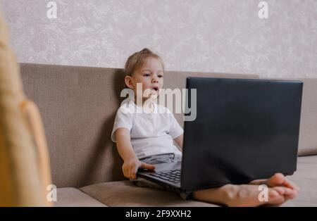 Ein kleines Mädchen sitzt zu Hause mit einem Laptop auf der Couch. Das Mädchen schaut während des Online-Unterrichts mit einem glücklichen Lächeln auf den Computerbildschirm. Home scho Stockfoto