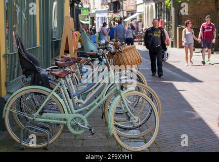 Staycation Idee. Eine Reihe von geparkten Fahrrädern vor Bells Fahrrädern und Menschen, die an einem Sommertag auf der George Street, Hastings Old Town, spazieren gehen. England. Stockfoto