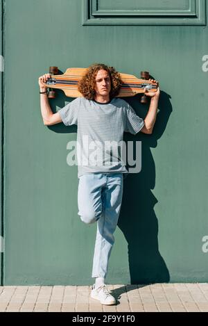 Attraktiver kaukasischer Sommersprossen-junger Mann, der Skateboard auf den Schultern hält. Er ruht auf einer grünen Mauer. Stockfoto