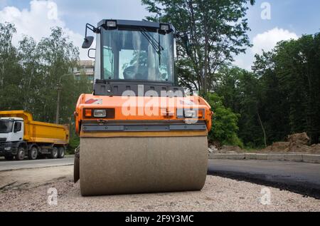 Große orangefarbene Walze auf Straßenbelag arbeitet. Im Hintergrund ist ein Muldenkipper. Verbesserung der Stadt. Ausrüstung für Straßenbau und Reparatur Stockfoto