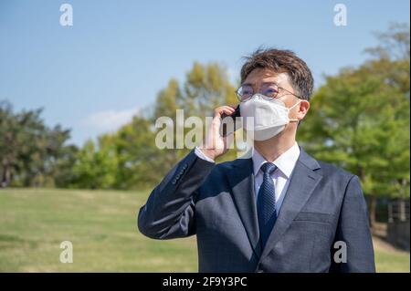 Ein asiatischer Geschäftsmann mittleren Alters, der eine Maske trägt und ein Smartphone auf dem Rasen verwendet. Stockfoto