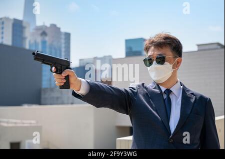 Ein asiatischer Mann, der eine Pistole in einer Sonnenbrille und einer Maske im Hintergrund der Stadt hält. Stockfoto