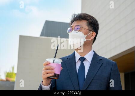 Ein asiatischer Geschäftsmann mittleren Alters in der Stadt, der eine Maske trägt und eine Kaffeetasse hält. Stockfoto