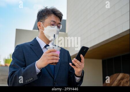 Ein asiatischer Geschäftsmann mittleren Alters in der Stadt, der eine Maske trägt, eine Kaffeetasse hält und ein Smartphone verwendet. Stockfoto