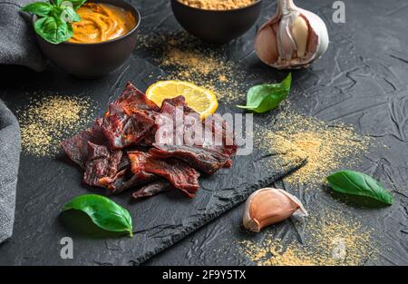 Dünne Scheiben getrocknetes Fleisch auf glänzendem schwarzen Hintergrund mit Gewürzen, Basilikum und Knoblauch. Seitenansicht mit Kopierbereich. Stockfoto