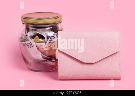Euro-Banknoten in Glas-Geldglas mit leerem Etikett und rosa Brieftasche, finanziell, sparen. Spardose mit leerem Haftnotizpapier. JAR voller Bargeld, sparen Stockfoto