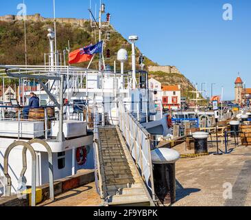 Ein Boot, das in einem Hafen mit einer Gangway nach oben vertäut ist. Eine Person sitzt auf dem Oberdeck und eine alte Burgmauer ist im Hintergrund. Stockfoto